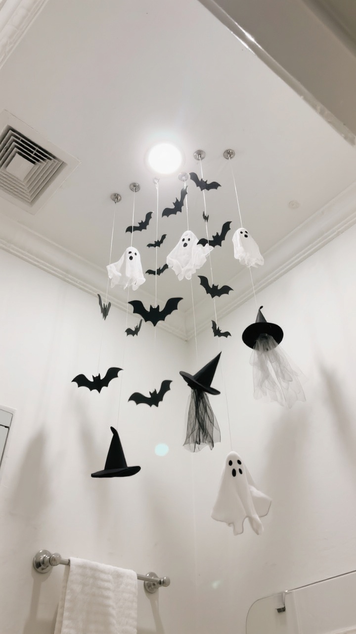 Upward-looking corner angle of a bathroom ceiling scene: suspended cluster of paper bats, tiny tulle ghosts, and a few mini witch hats hanging from clear fishing line at varied heights for a swarm effect; discreet Command hooks visible upon close inspection; clean white ceiling with crown molding; keep clear of vent fan area; subtle shadow play from ambient ceiling light for immersive movement, no other decor distractions.